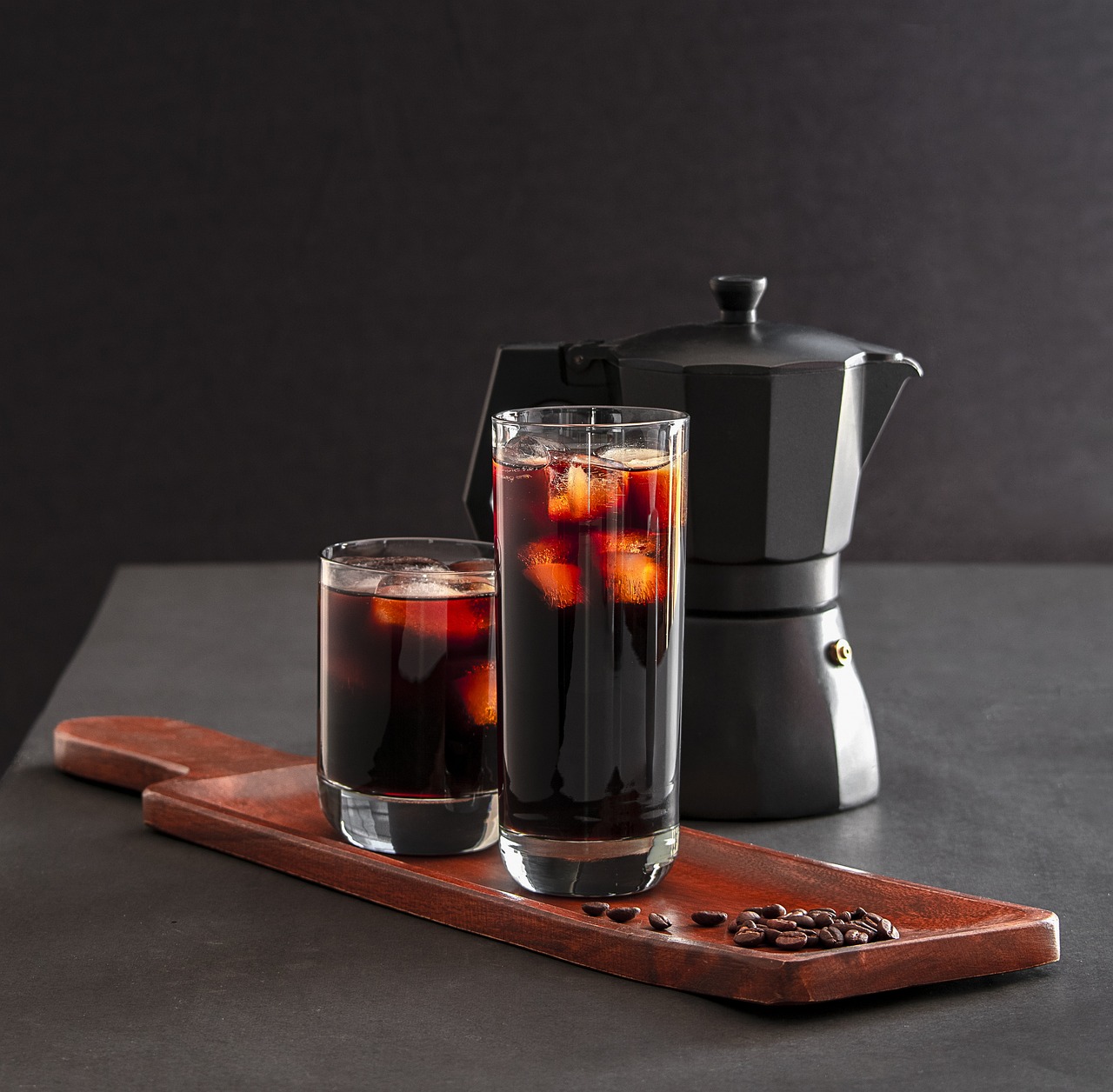 Cold brew coffee and iced coffee in clear glasses alongside a classic coffee moka pot, displayed on a wooden board with scattered coffee beans for a rustic presentation.